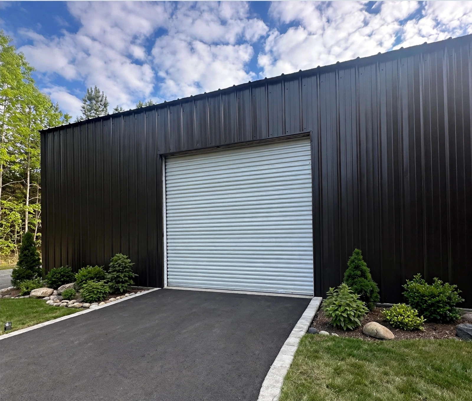 Finished black metal building with a white commercial roll-up door, paved driveway and landscaped planting beds
